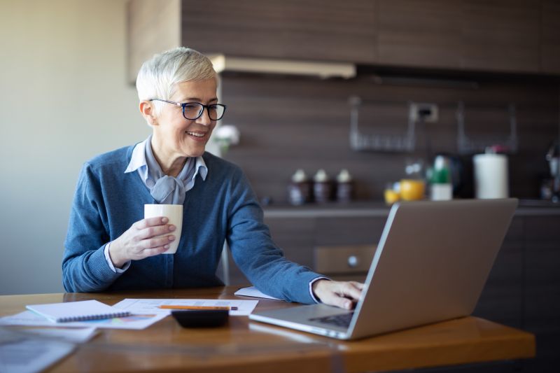An older man sitting at a table, drinking a cup of coffee with one hand on his laptop